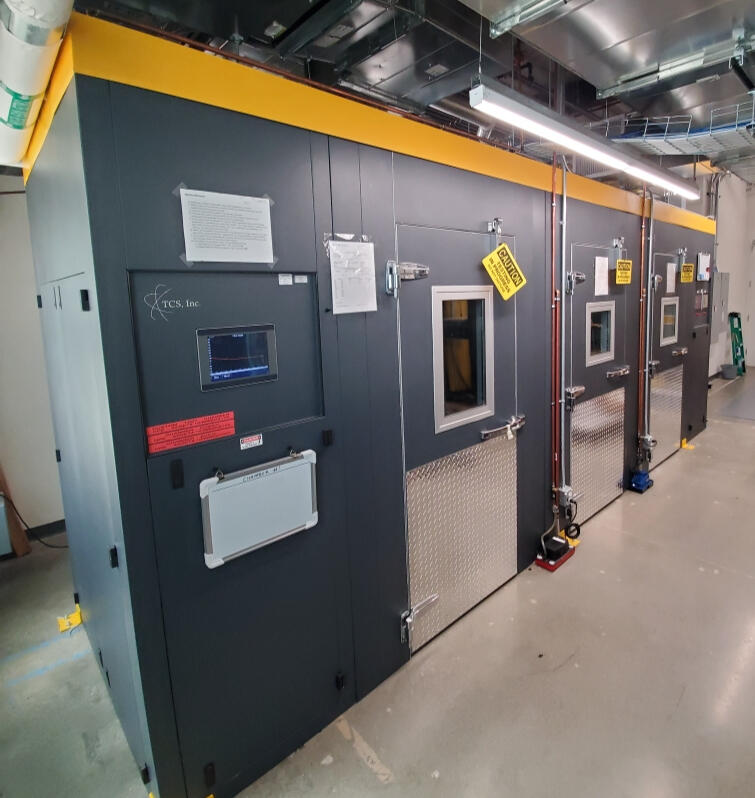 Custom Walk-In Environmental Test Chambers by TCS, Inc. A row of custom-built walk-in environmental test chambers with durable gray and yellow finishes, featuring diamond-plated doors and advanced control panels for precision testing in a high-performance lab setting.